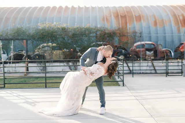 Groom dipping the bride into a kiss out on the concrete courtyard, with the hedge, the parked old cars and the curved metal roof of the building behind them