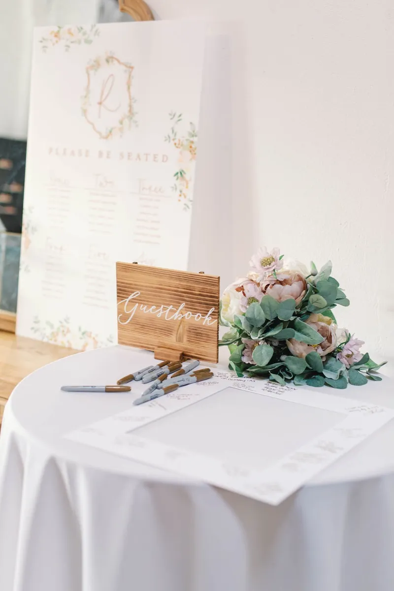 Guest book table with a small wooden sign, a row of pens laid out and a bouquet of blush flowers and eucalyptus beside the seating chart
