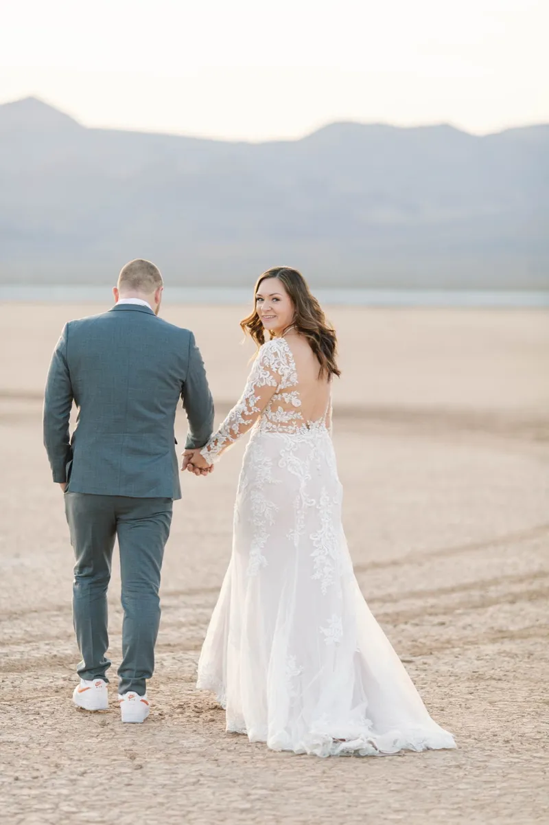 A couple walking across the dry lake bed at sunset while the bride looks back over her shoulder