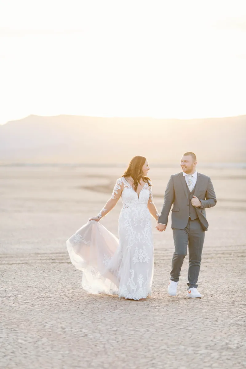 Bride in a long sleeve lace gown holding her train and walking with her groom into the sun