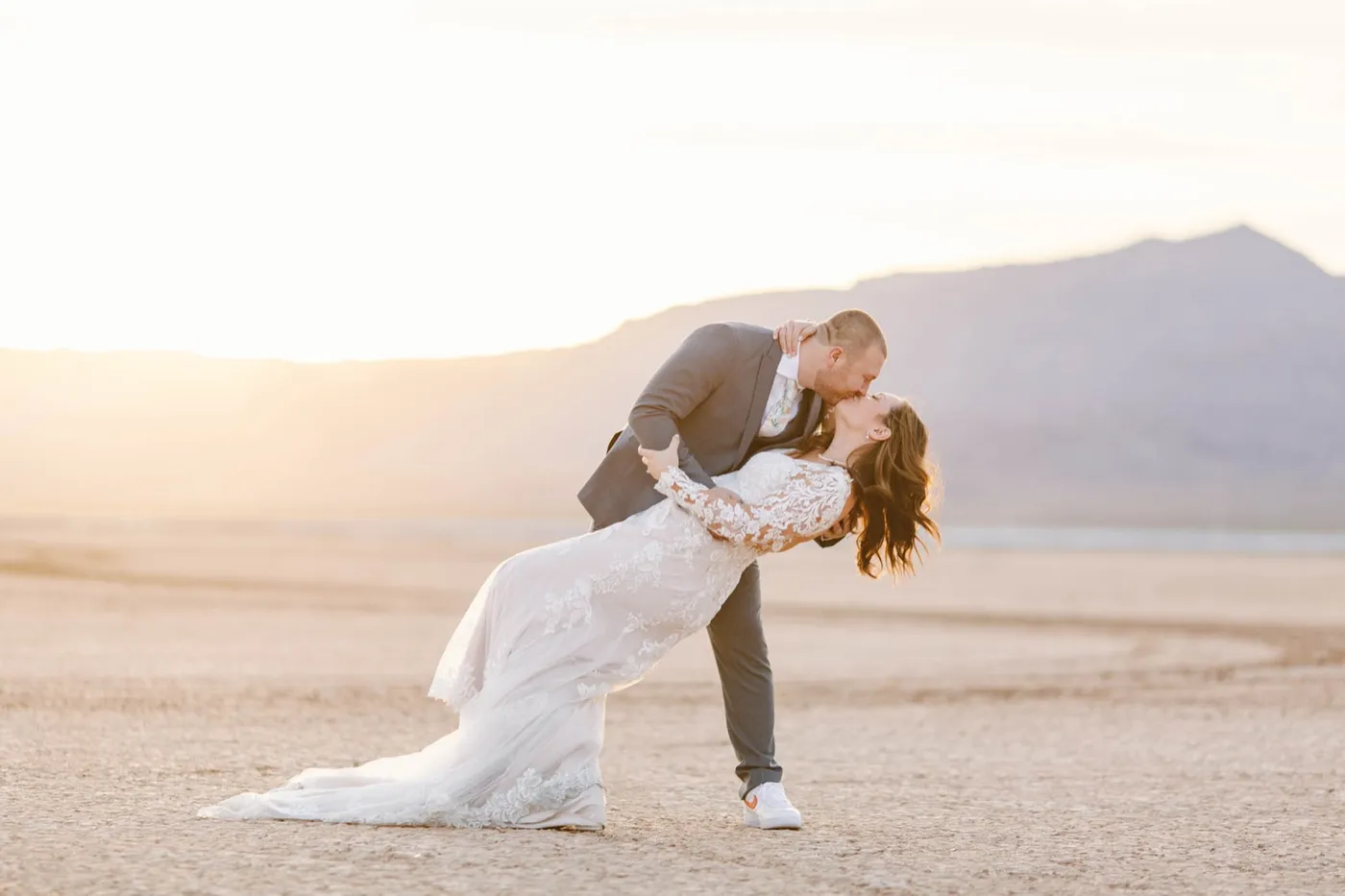 Groom in a grey suit dipping his bride for a kiss at sunset, her lace train sweeping the cracked dry lake bed