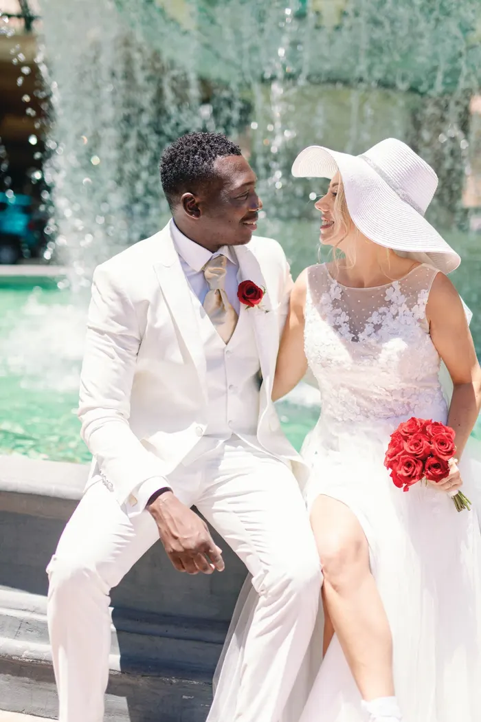 A groom in a white suit and his bride in a sun hat celebrating at a Las Vegas fountain