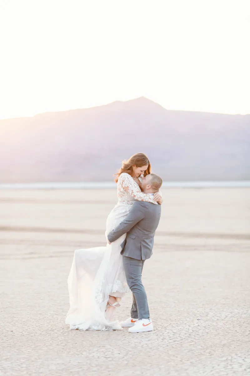 Groom in a grey suit lifting his bride off the ground on the lake bed as the sun sets behind them