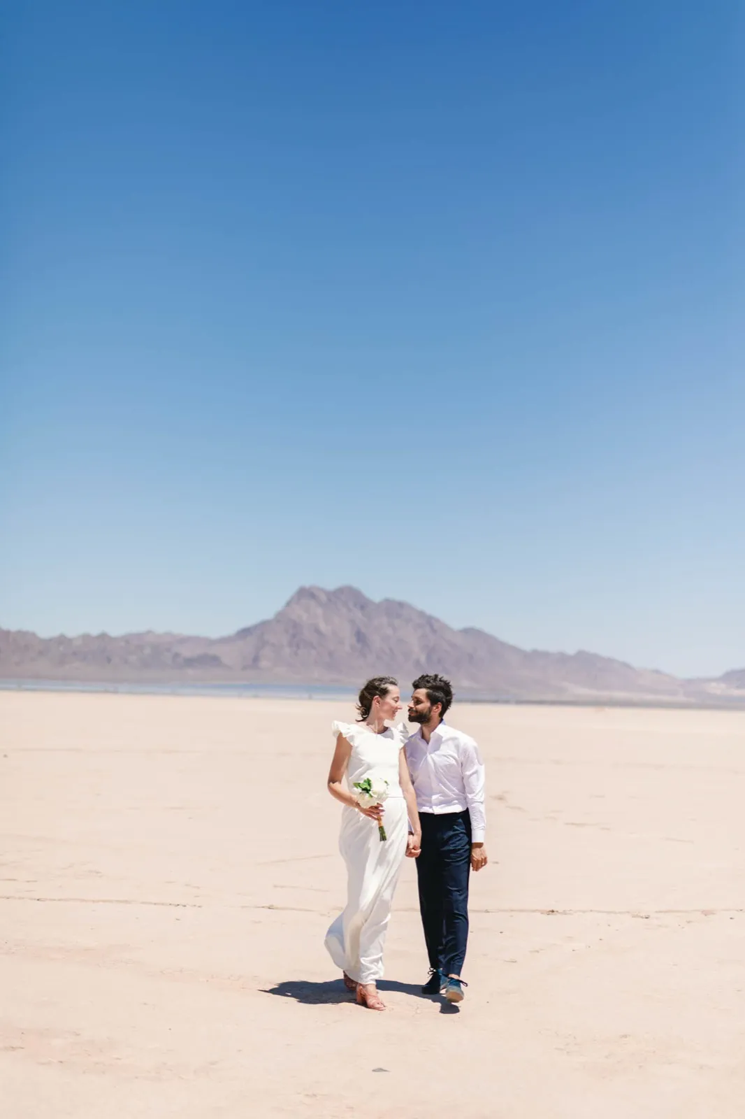 Bride and groom walking hand in hand across a pale cracked lake bed with a single dark peak on the horizon