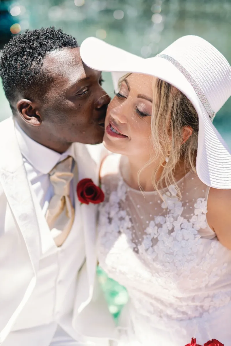 A groom kissing the cheek of his bride in a wide white sun hat beside a Las Vegas fountain