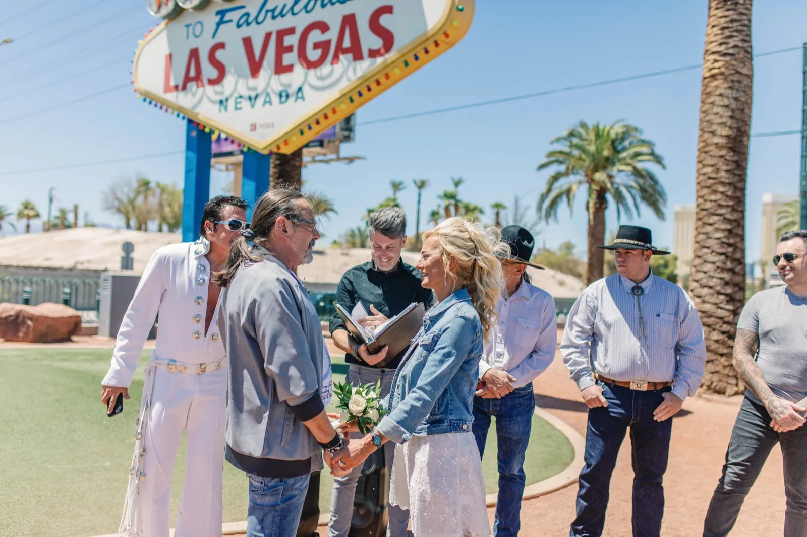 Officiant reads from a folder as a couple hold hands under the Las Vegas sign, guests in cowboy hats watching