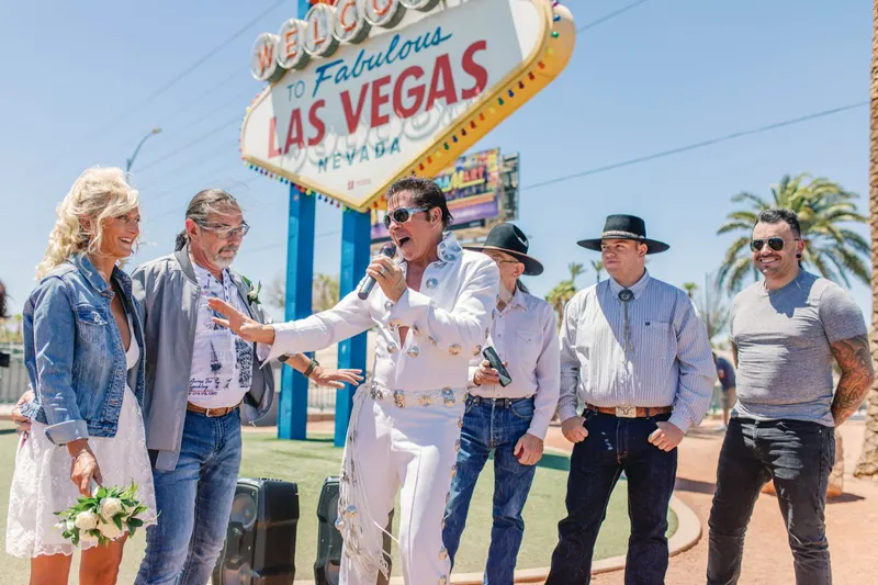 Elvis impersonator in a white jumpsuit sings under the Las Vegas sign as the bride, groom and guests in cowboy hats watch