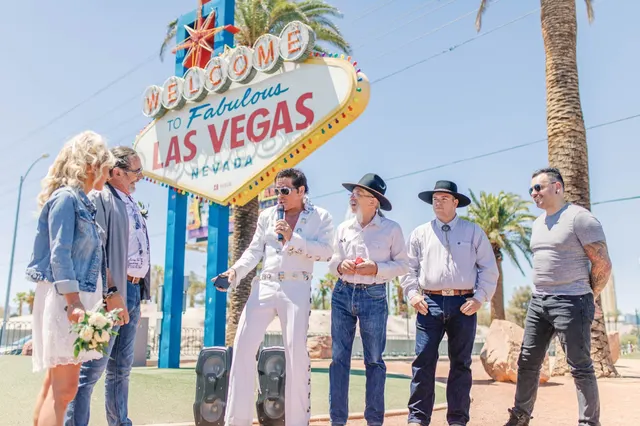 Bride and groom standing to one side while an Elvis impersonator sings into a microphone under the Welcome to Fabulous Las Vegas sign