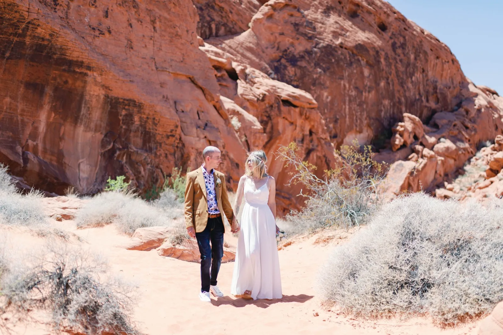 Bride and groom holding hands as they walk across pink sand below a towering wall of red sandstone at Valley of Fire