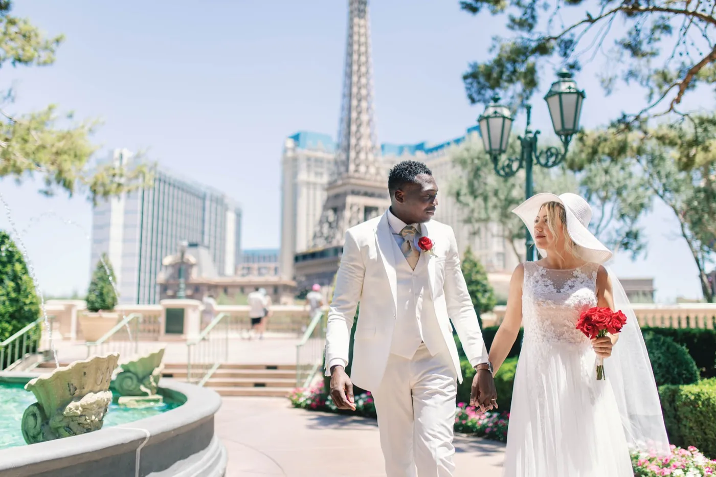Groom in a white suit and bride in a wide-brim hat walking hand in hand past a Bellagio fountain, Paris tower behind