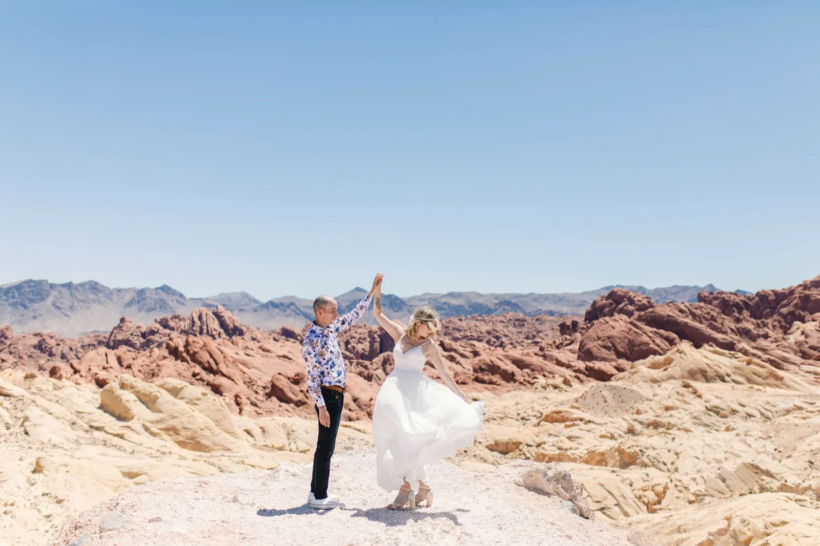 Groom in a blue floral shirt lifts his bride’s hand overhead on bare white sandstone at Valley of Fire
