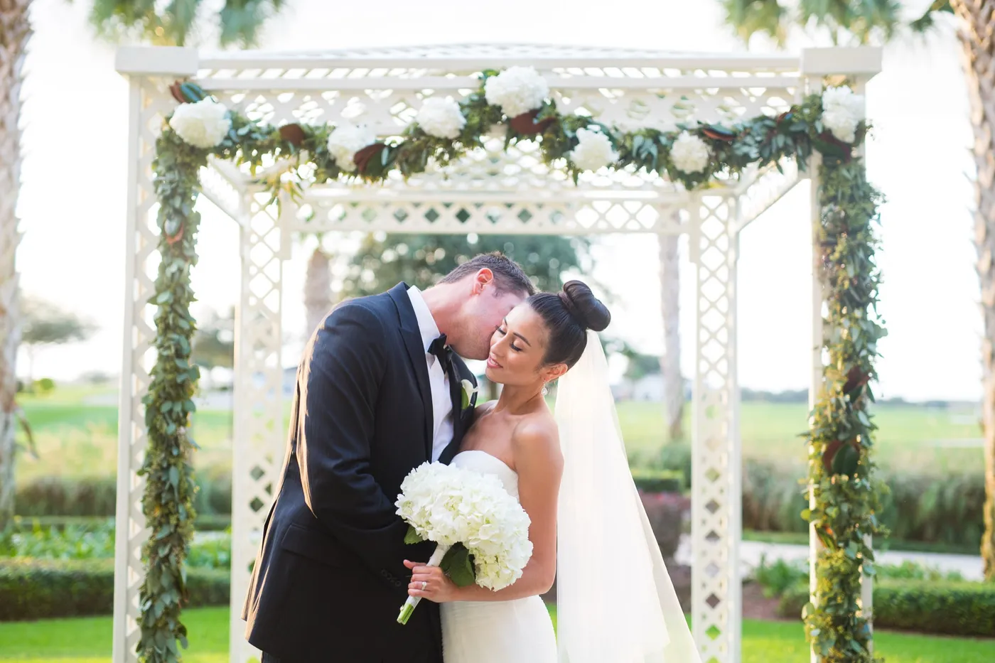Groom in a black tuxedo kissing his bride's cheek under a white arbor draped with hydrangeas and eucalyptus