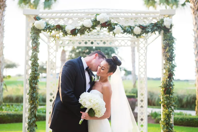 Groom in a black tuxedo kissing his bride's cheek under a white arbor draped with hydrangeas and eucalyptus