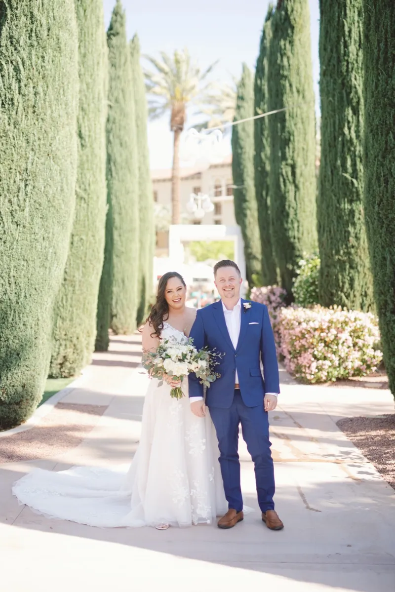 Bride and groom standing arm in arm on the paved walk between tall cypress trees with the resort behind them