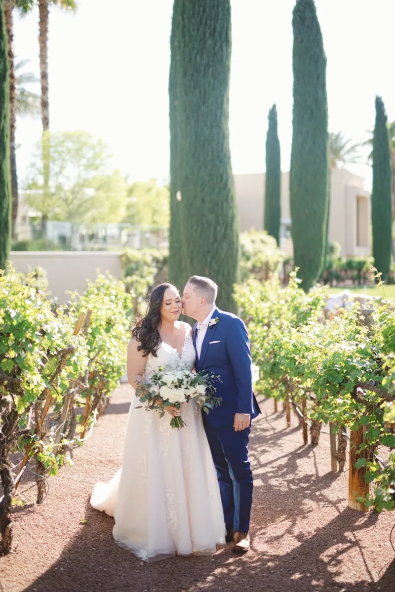 Groom kissing the bride's head as they stand on the gravel path between the grapevine rows