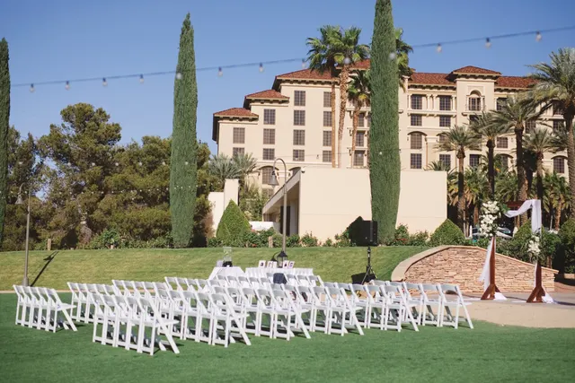 Rows of white chairs on the lawn facing a floral arch, with the resort's tan towers, palms and cypress trees behind