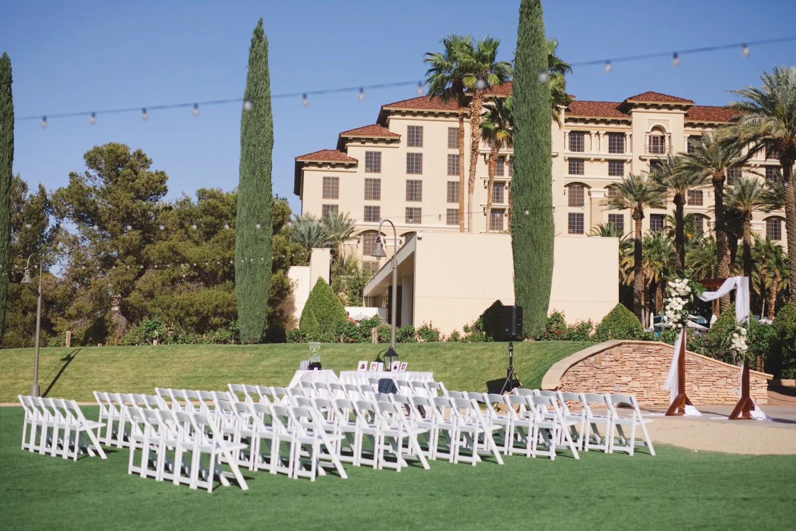 Rows of white folding chairs face a floral arch on a green lawn below the resort’s tan towers and palm trees