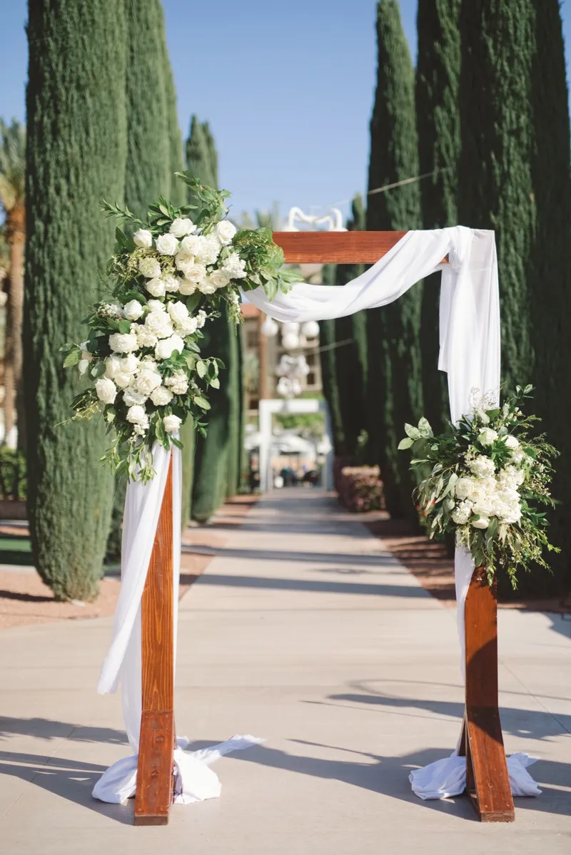 Wooden ceremony arch with white drape and white flowers standing at the head of the aisle between cypress trees