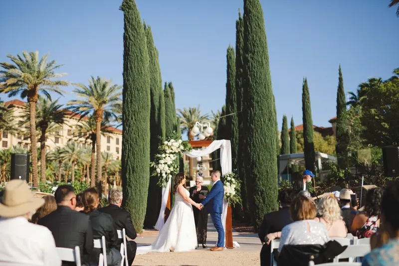 A bride and groom hold hands under a draped wooden arch on a lawn as seated guests watch, tall cypress trees behind