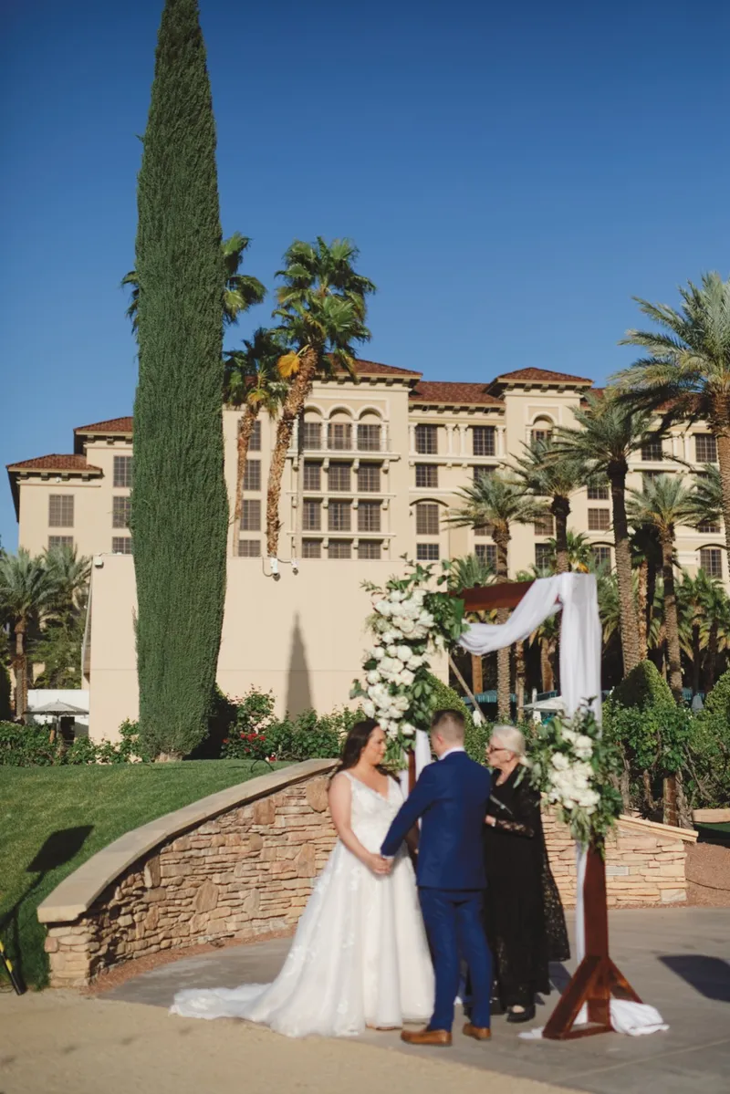 Bride and groom standing hand in hand with their officiant at the flower topped arch, the resort building rising behind