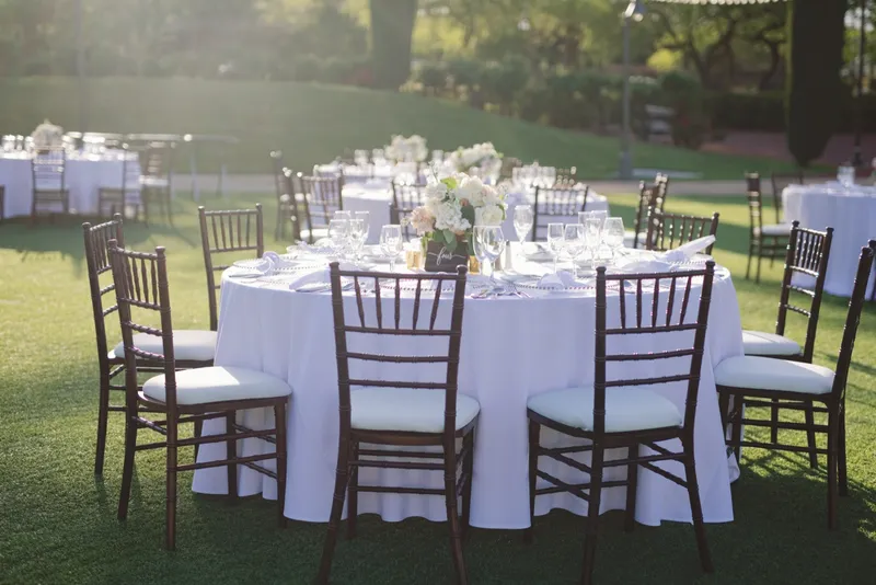 A round reception table set on the grass with white linen, wooden chairs and a low white centerpiece