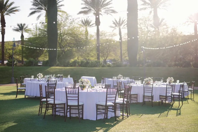 Wide view of the lawn reception at sunset, round tables under cafe lights with palm trees behind