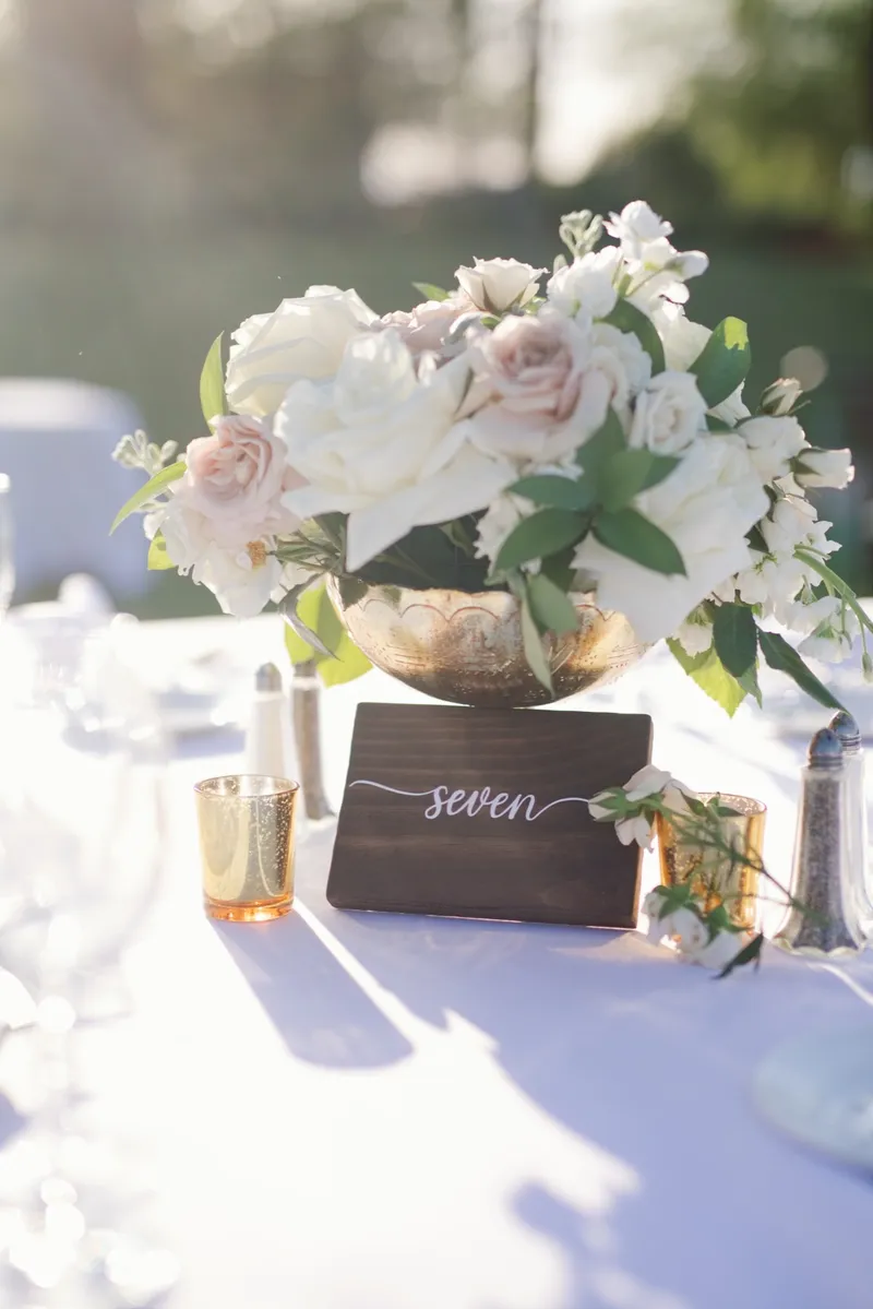 A wooden table number seven beside a footed gold bowl of white and blush flowers