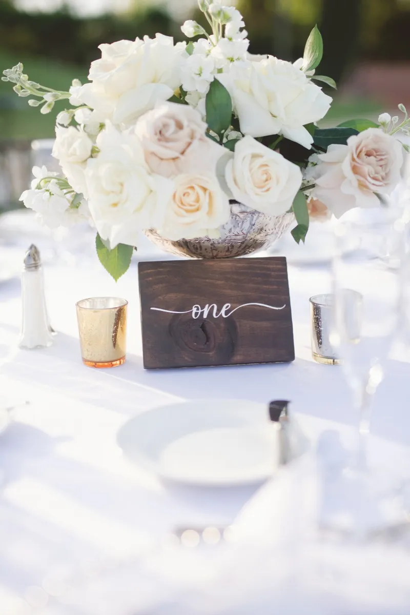 A wooden table number one at a set place with white roses in a footed bowl above it