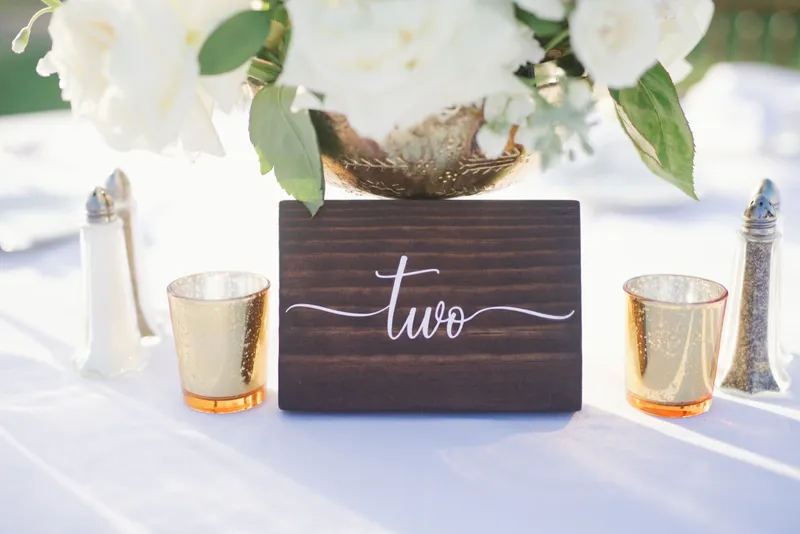 A wooden table number two standing between gold votives with white flowers above it