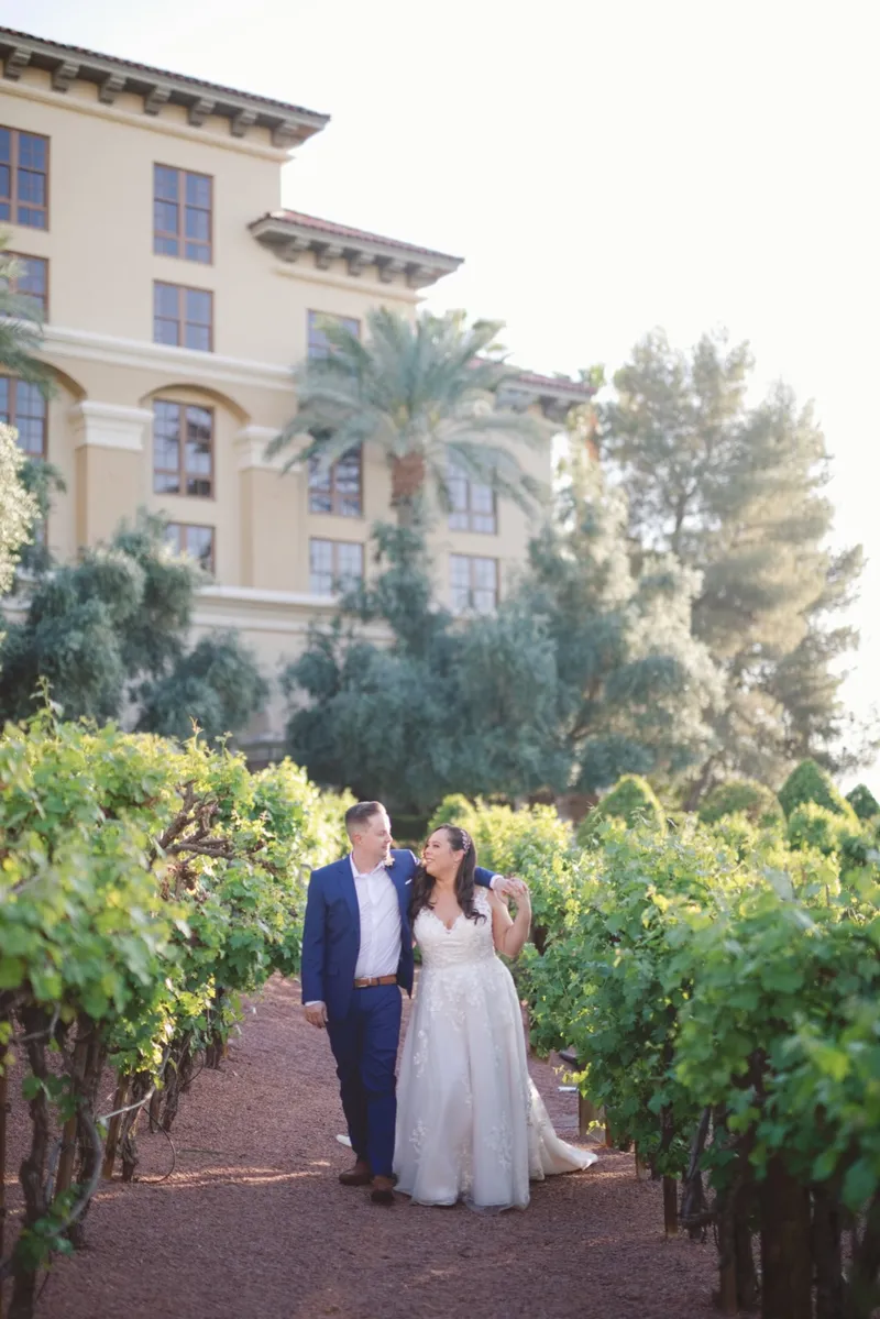 Bride and groom walking arm in arm down the gravel path between grapevine rows, the resort tower behind them