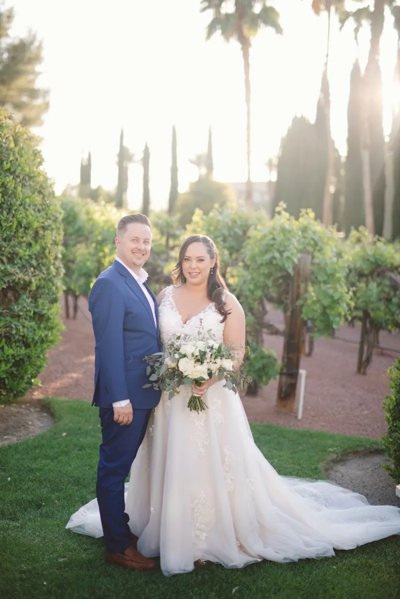 Bride and groom standing together at the end of the vineyard rows as the sun flares through the palms behind them