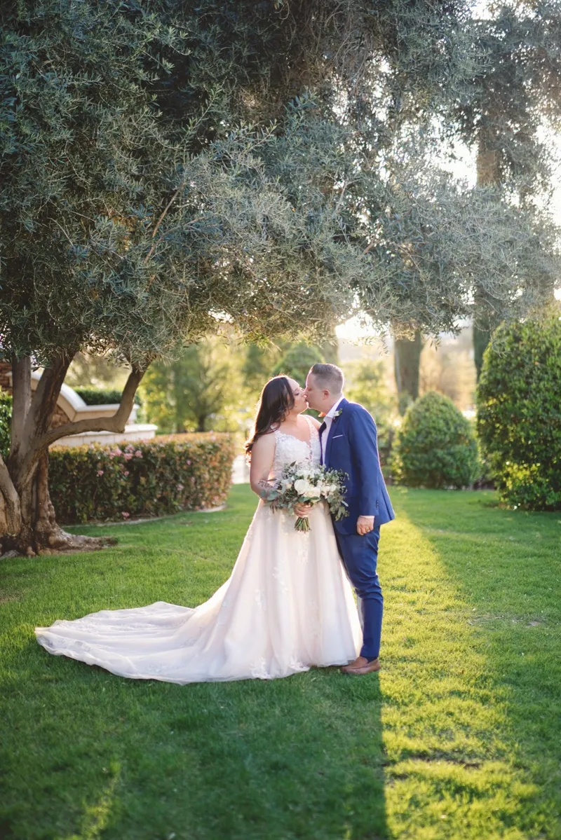 Bride and groom kissing on the lawn beneath a spreading olive tree, her train laid out on the grass