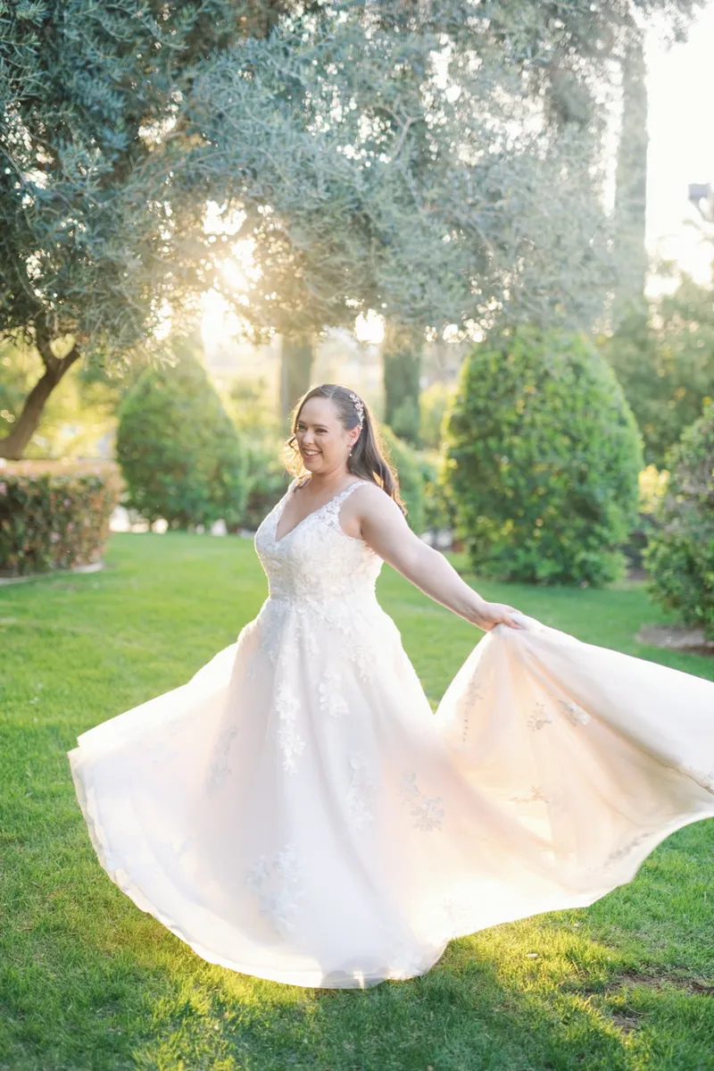 Bride swinging her skirt out wide with both hands, smiling, the sun coming through the olive tree behind her