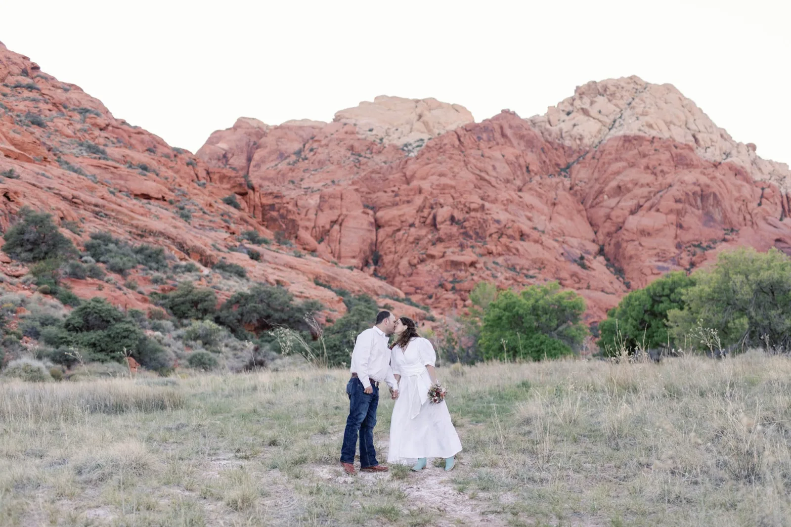 Bride and groom kissing in a grassy wash below the red sandstone cliffs of Red Rock Canyon after their elopement