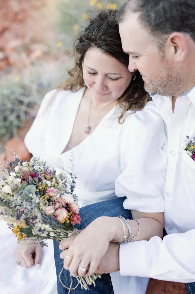 Couple sitting close together in the brush with her dried bouquet across her lap