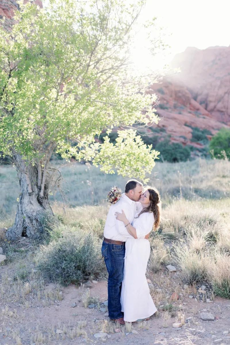 Couple kissing under a pale desert tree with the red cliffs beyond