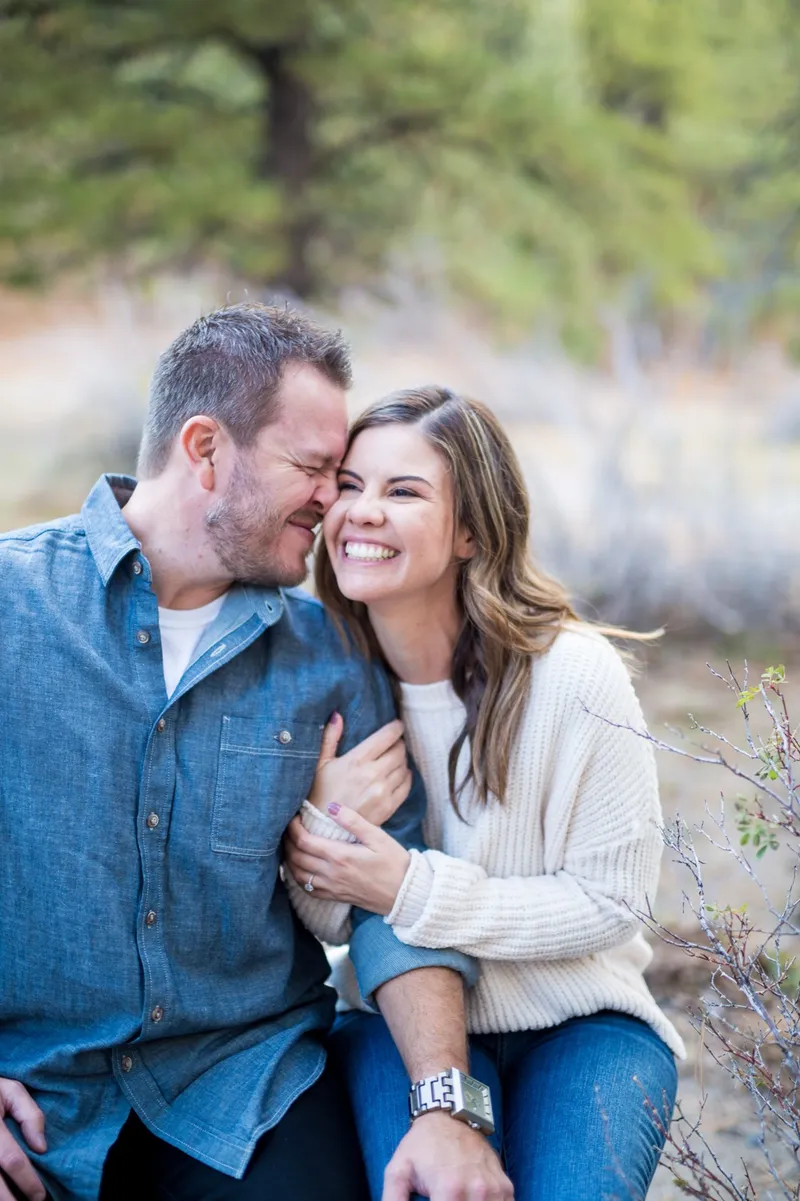 Man in a chambray shirt laughing with his fiancee in a cream sweater, their foreheads touching