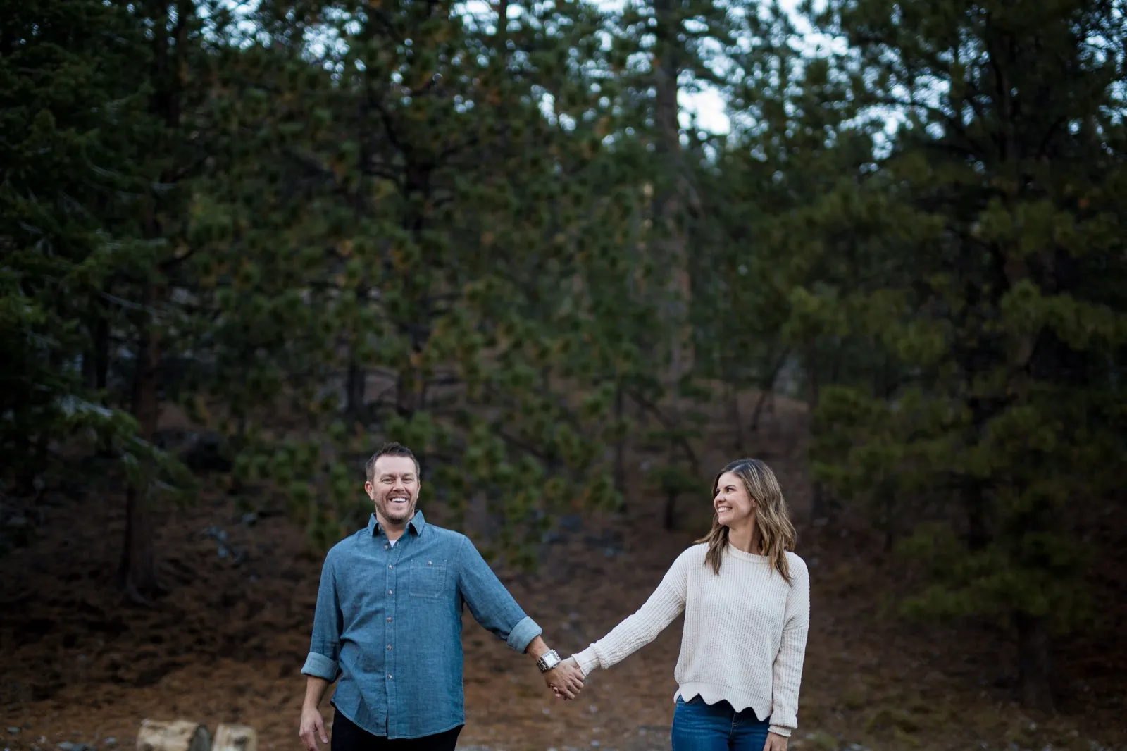 Couple walking toward the camera holding hands across a pine needle clearing in front of tall shaded pines