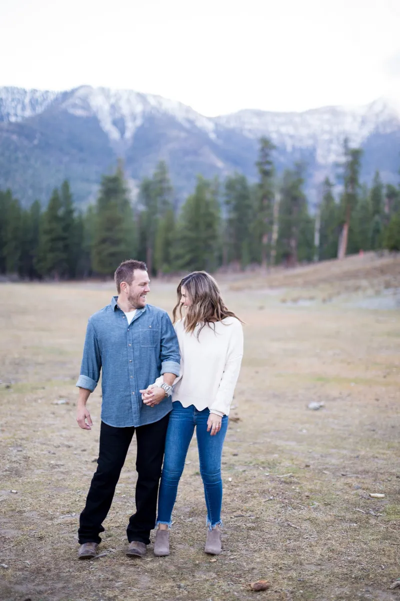 Couple standing in a dry meadow holding hands with a long snow covered ridge behind the pines