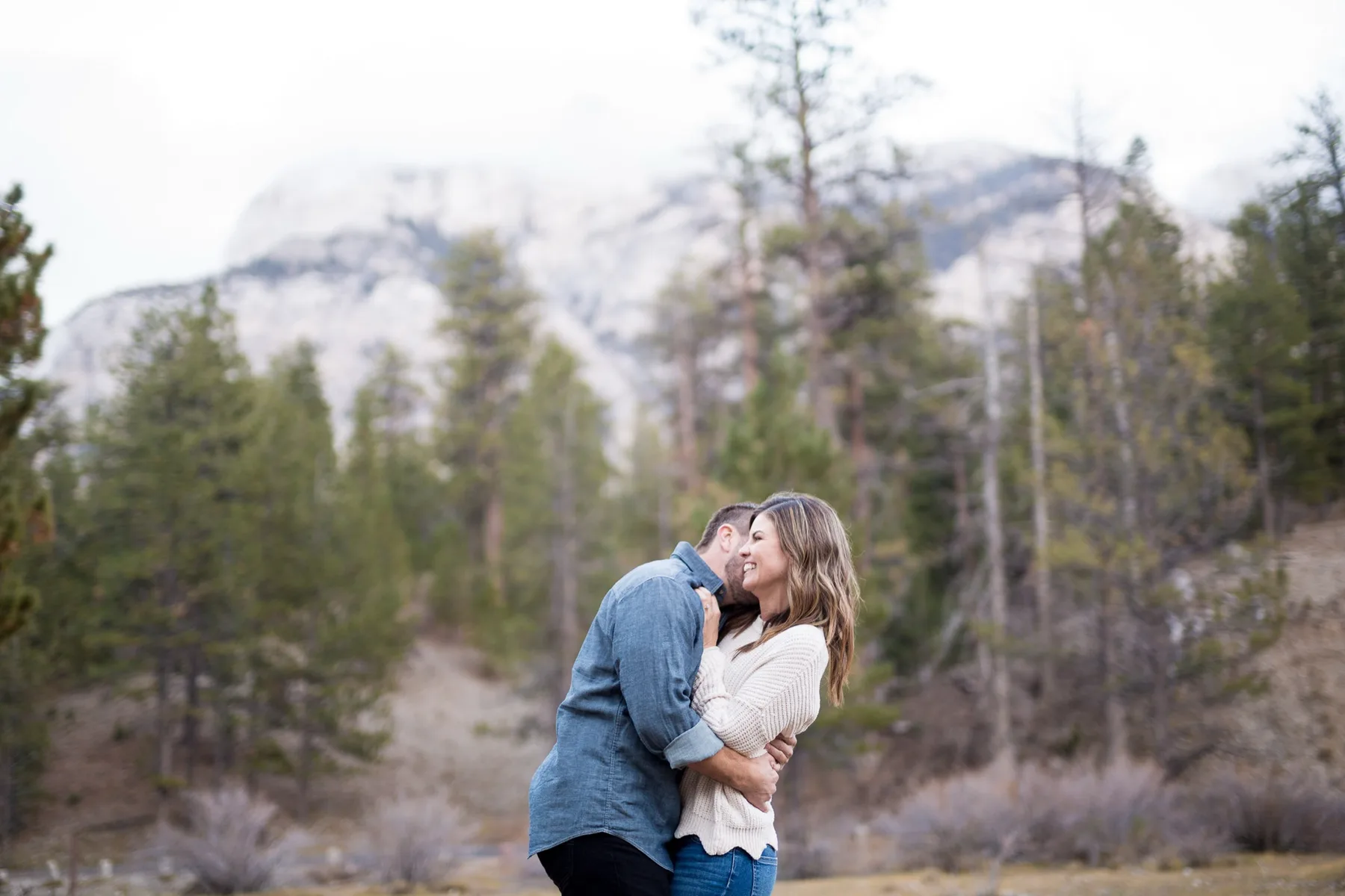 Couple kissing in the pines at Mount Charleston with snowy peaks behind them