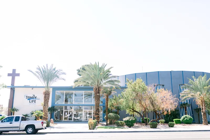 The church seen from across the street, a low blue and white building behind a row of palms with a cross standing near the front doors