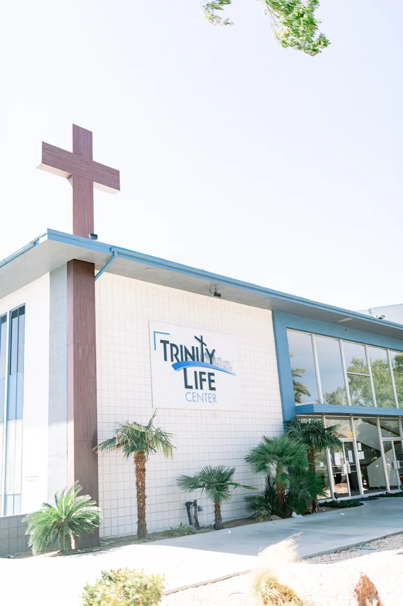 The corner of the church building with the Trinity Life Center sign on the wall and a tall cross mounted on the roofline against a clear sky