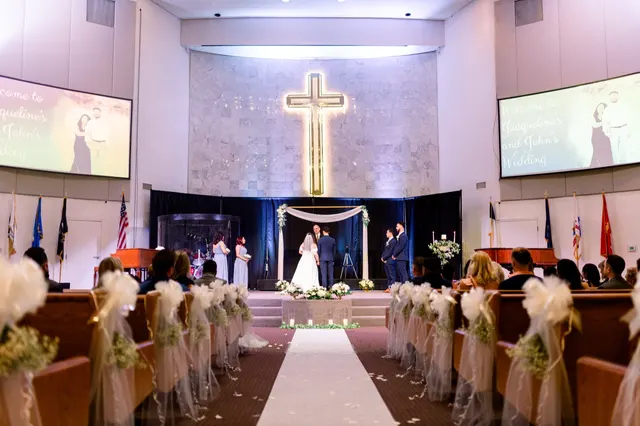 Wedding ceremony seen from the back of the sanctuary, the couple standing under a white draped arch with a tall lit cross on the pale stone wall above them and pews dressed in tulle down both sides of the aisle