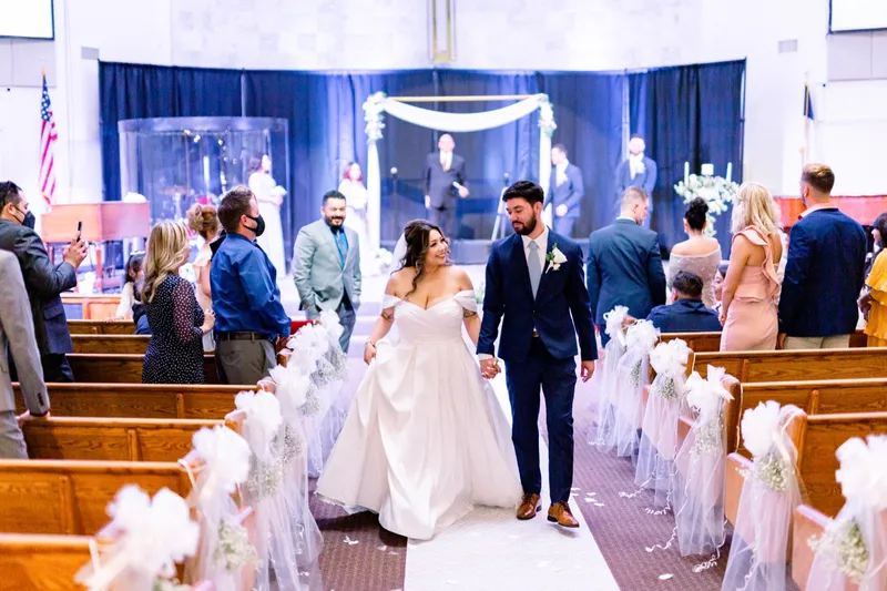 The recessional from the back of the church, the couple walking hand in hand past guests standing in the pews with petals on the white runner behind them