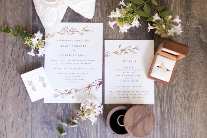 Invitation suite laid out on weathered wood with white blossoms, drop earrings and two open ring boxes beside it