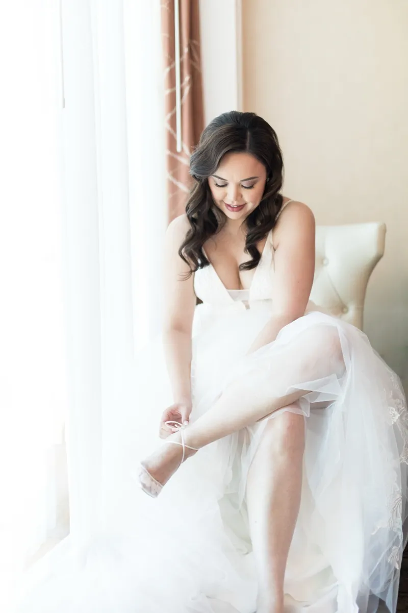 Bride sitting in window light fastening her shoe with her tulle skirt spread around her