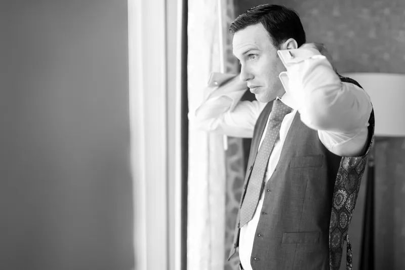 Black and white frame of the groom straightening his collar and tie at the window