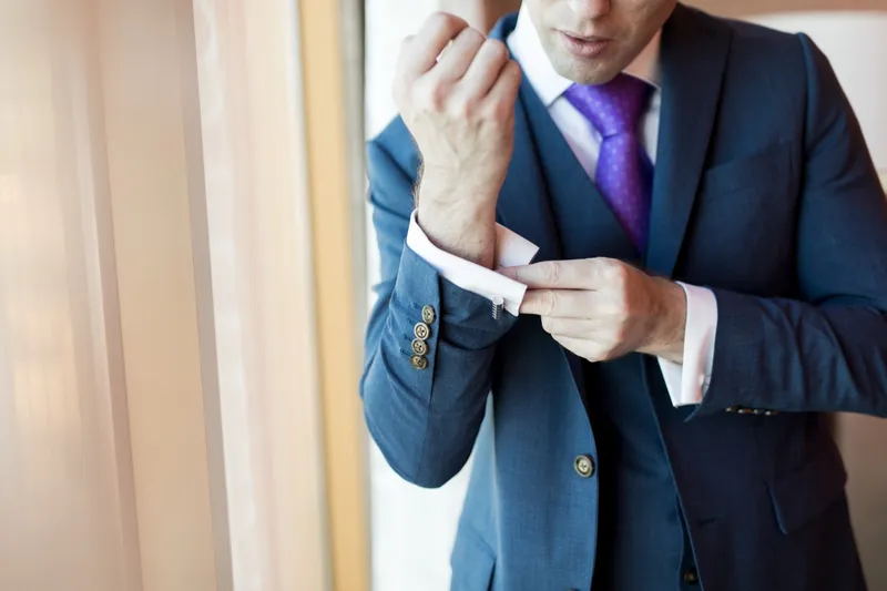 Groom fastening a cufflink in a blue suit and purple tie by the window