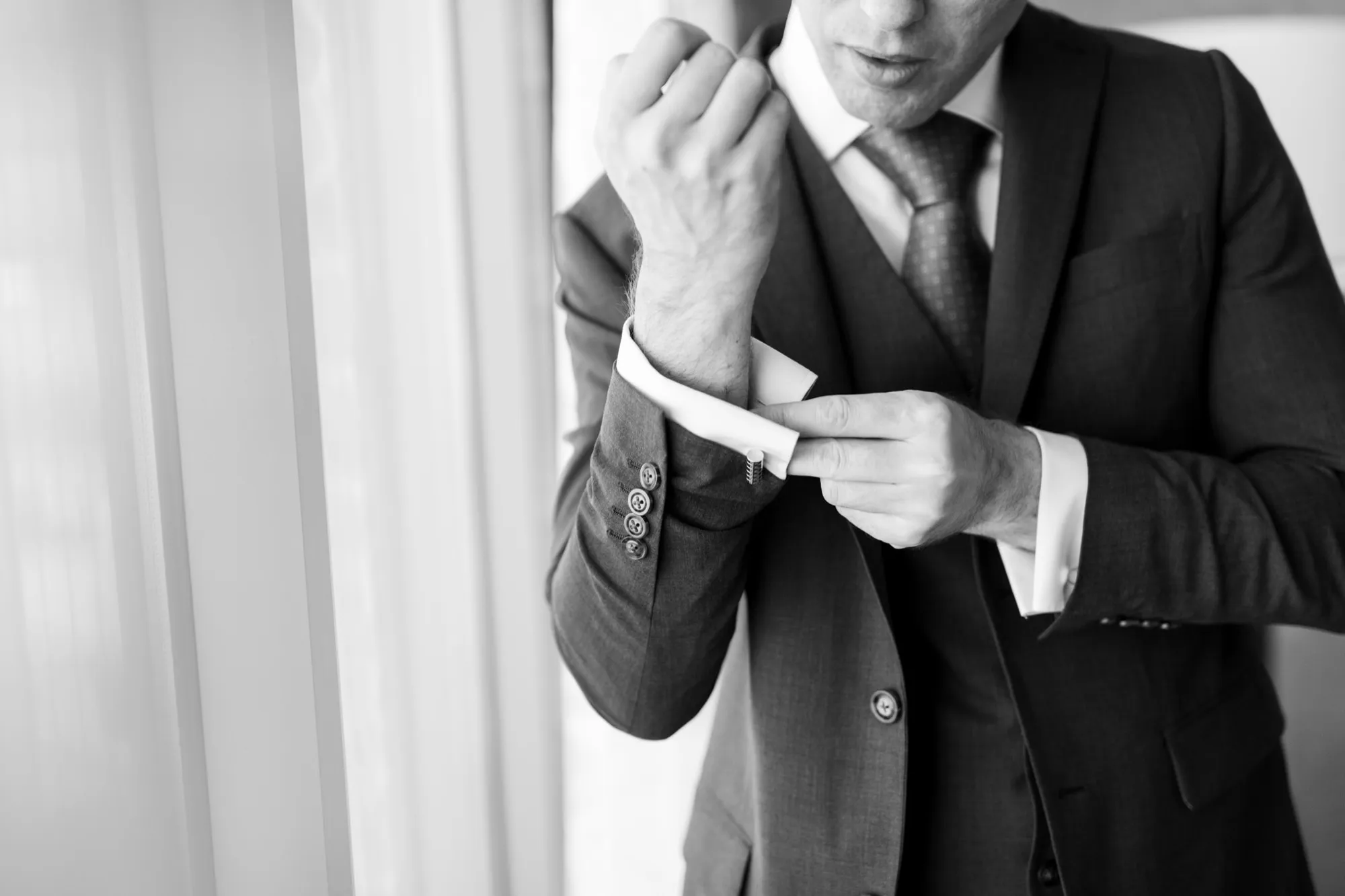 Black and white photo of a groom fastening his cuff link by a window while he dresses