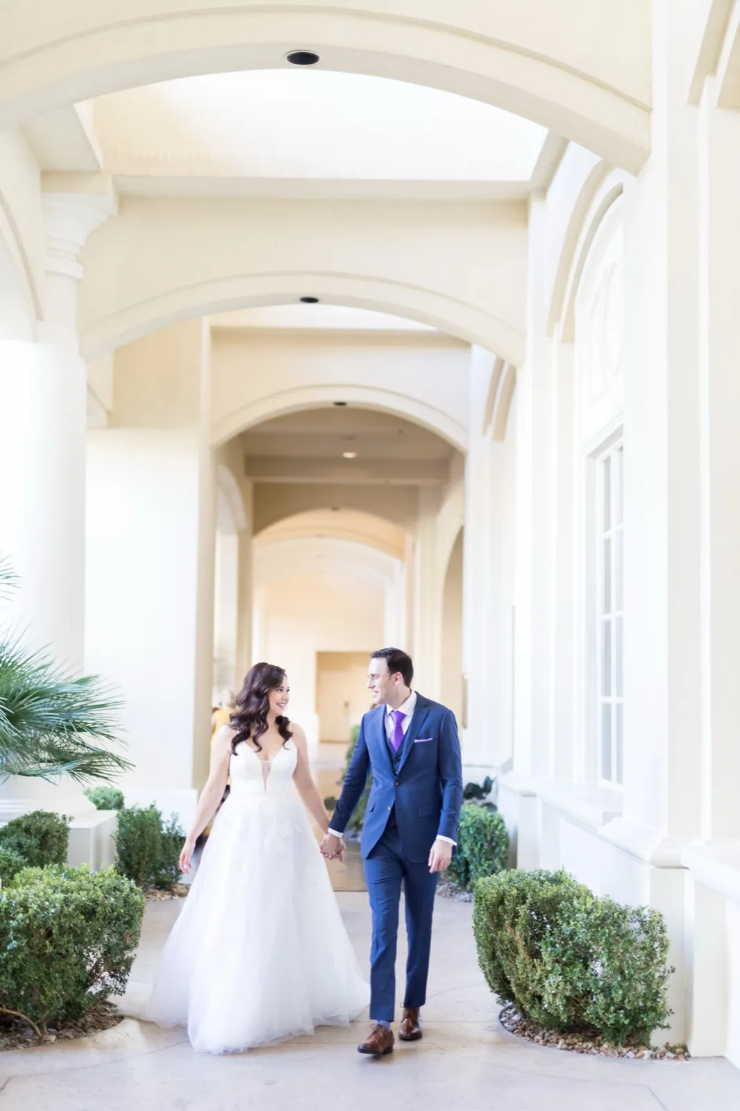 Bride and groom hold hands walking a sunlit arched colonnade lined with clipped hedges and a palm